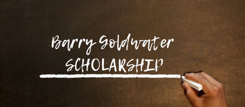 Hand writing the words "Barry Goldwater Scholarship" on a chalkboard with a piece of white chalk.
