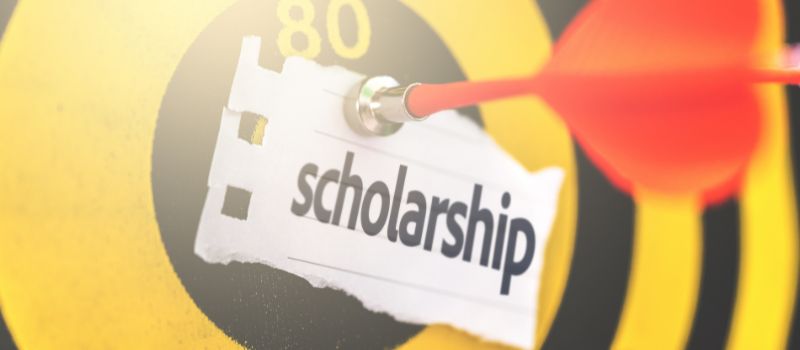 A close-up of a dartboard with a dart hitting the bullseye, and a piece of paper with the word "scholarship" written on it.
