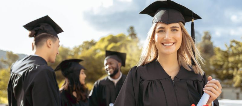 Graduates in black caps and gowns with one holding a diploma, standing outdoors with trees and a bright sky in the background.
