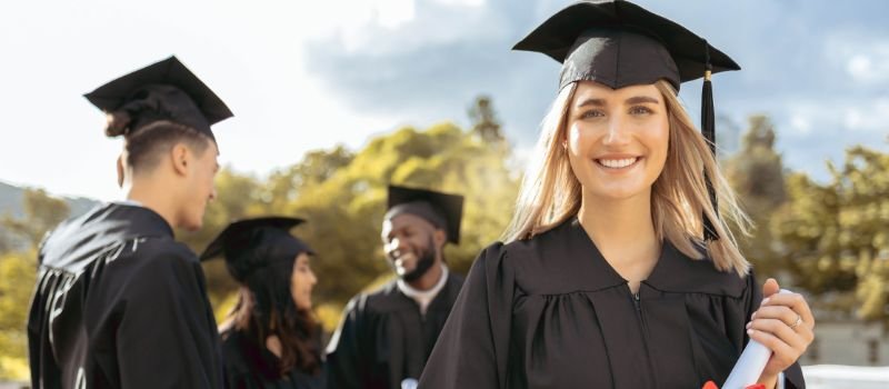Graduates in black caps and gowns with one holding a diploma, standing outdoors with trees and a bright sky in the background.