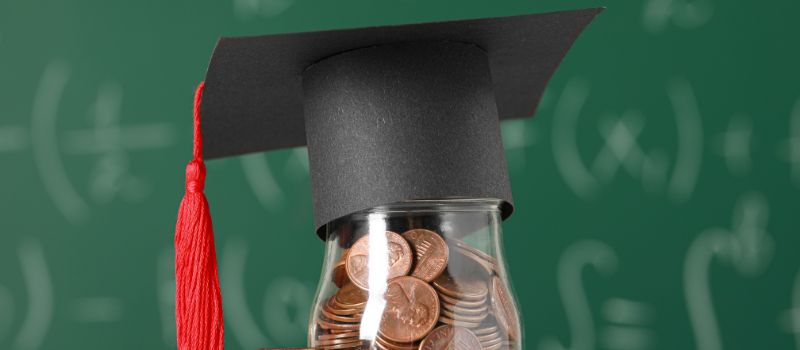 A graduation cap resting on top of a glass jar filled with coins, with a green chalkboard in the background featuring mathematical equations.

