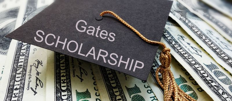 A graduation cap with "Gates Scholarship" printed on it, resting on top of a pile of dollar bills.
