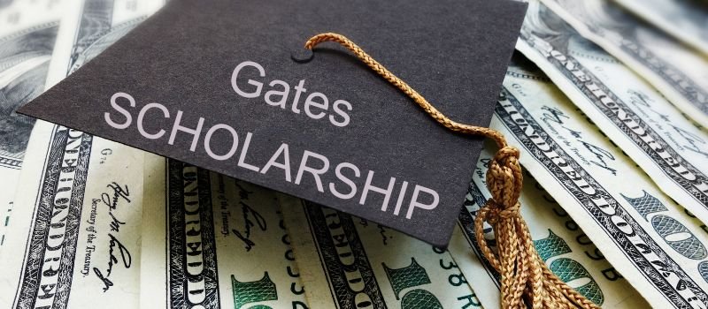 A graduation cap with "Gates Scholarship" printed on it, resting on top of a pile of dollar bills.