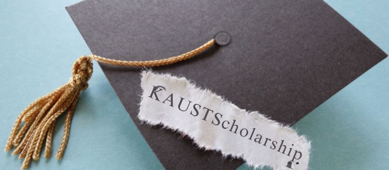 A black graduation cap with a gold tassel on a light blue background, with a piece of torn white paper with "KAUSTscholarship" printed on it.
