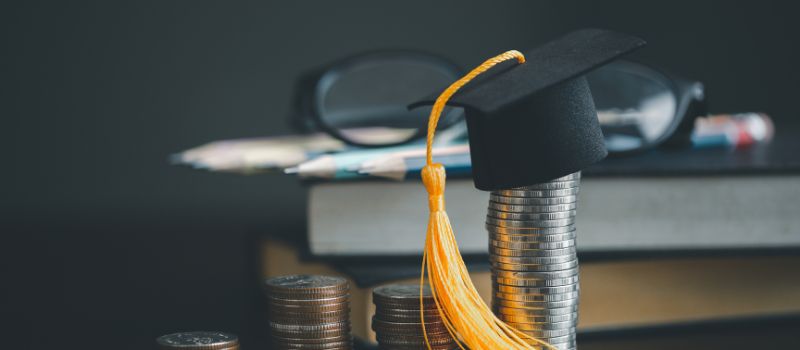 Black graduation cap with a yellow tassel sitting on top of a stack of coins next to more stacks of coins and books with a magnifying glass in the background.
