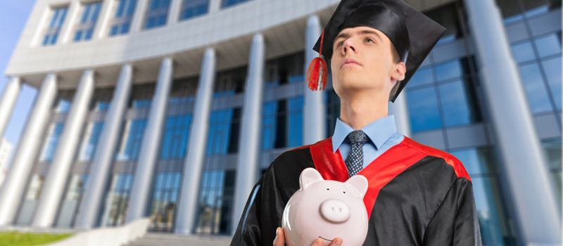A graduate in a black and red academic gown and mortarboard holding a piggy bank in front of a modern university building.
