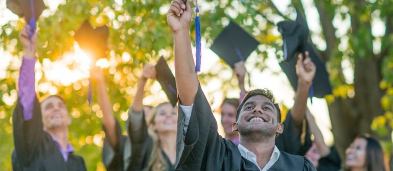 Graduates in black gowns and caps joyfully raising their hands and diplomas into the air, with a sunlit backdrop of green trees.