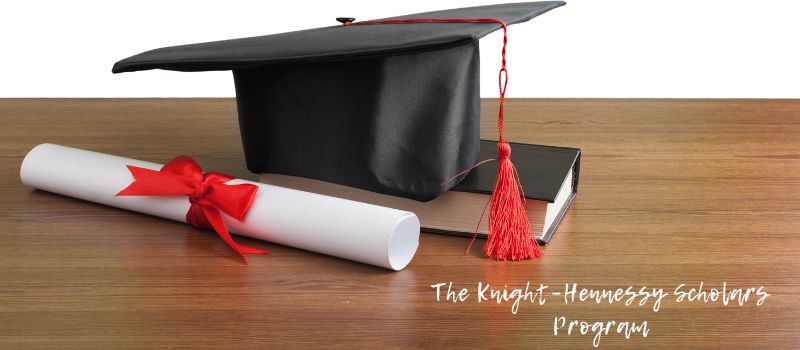 A black graduation cap with a red tassel sitting on top of a book, next to a rolled diploma tied with a red ribbon, with text "The Knight-Hennessy Scholars Program" in the bottom right corner.

